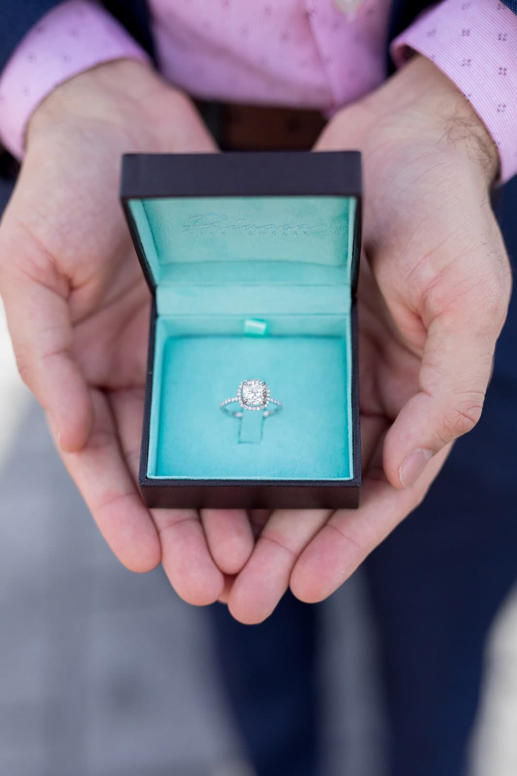 A Privosa cushion halo engagement ring in its box, held in two hands